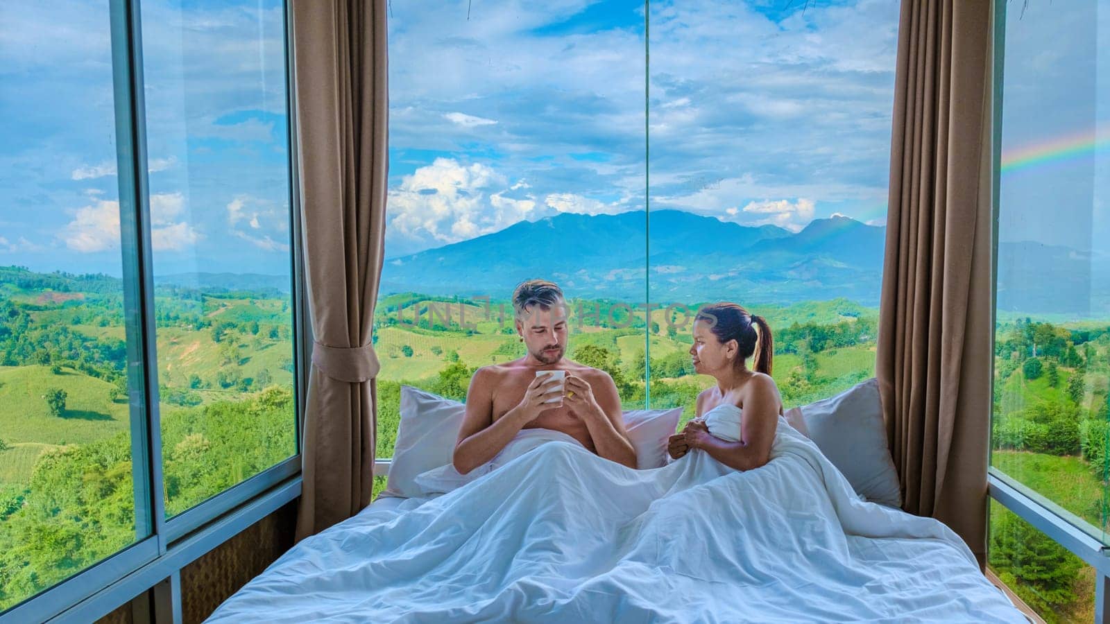 a couple of men and women laying in bed with open widows looking out over the mountains of Thailand, men and woman in bedroom with large open glass windows looking out over the green landscape of Nan
