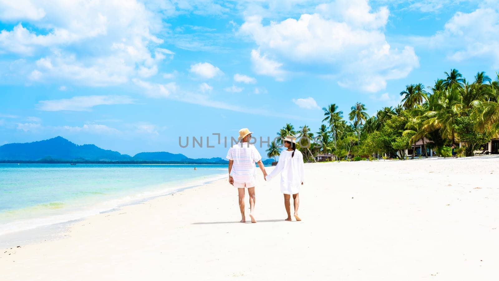 a couple of men and woman with summer Koh Muk tropical island, a caucasian man and Asian woman walking on a white beach with palm trees during summer holiday vacation in Koh Mook Trang Thailand