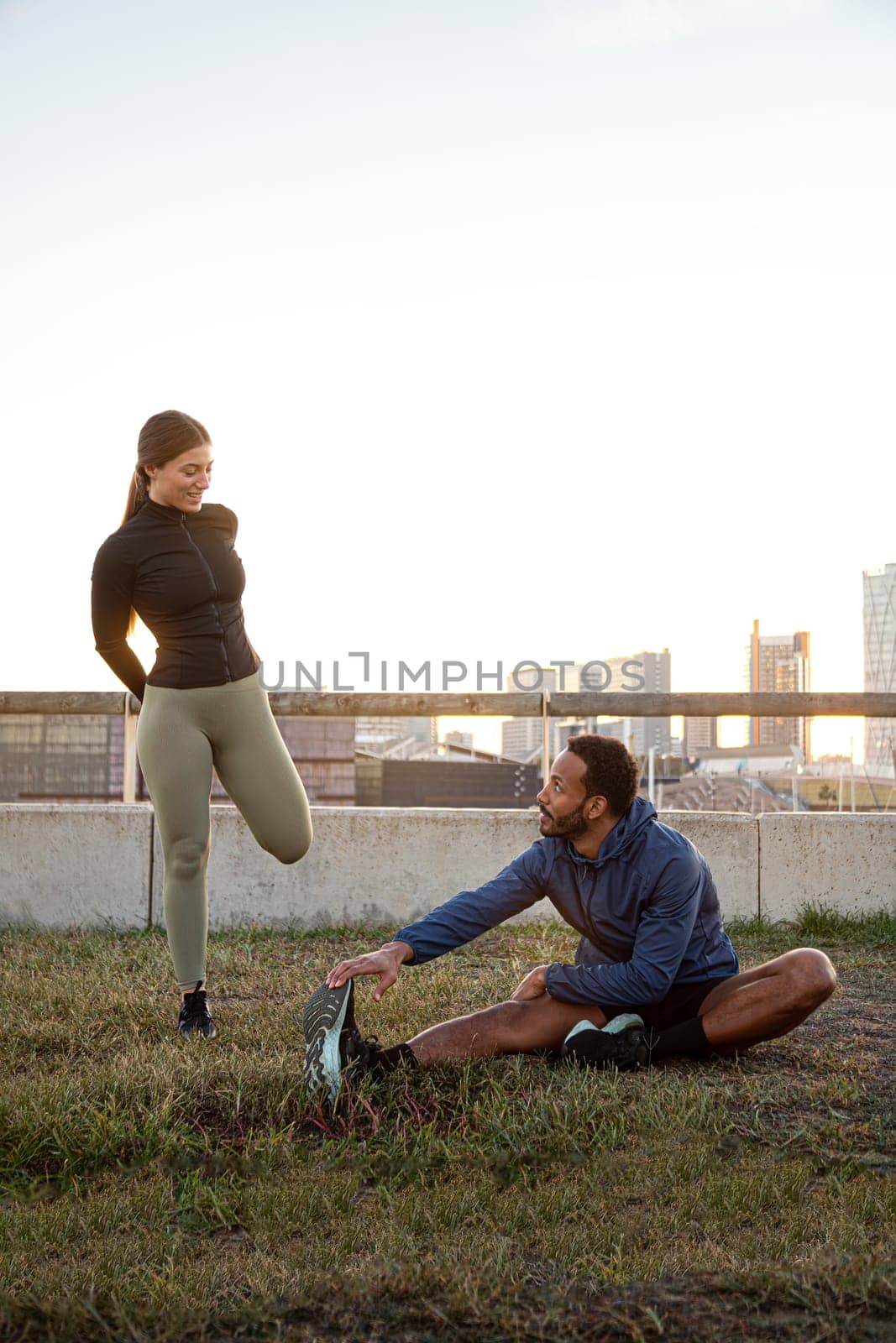 Vertical portrait of young multiracial couple stretching leg before running, training outdoors. Copy space. by Hoverstock