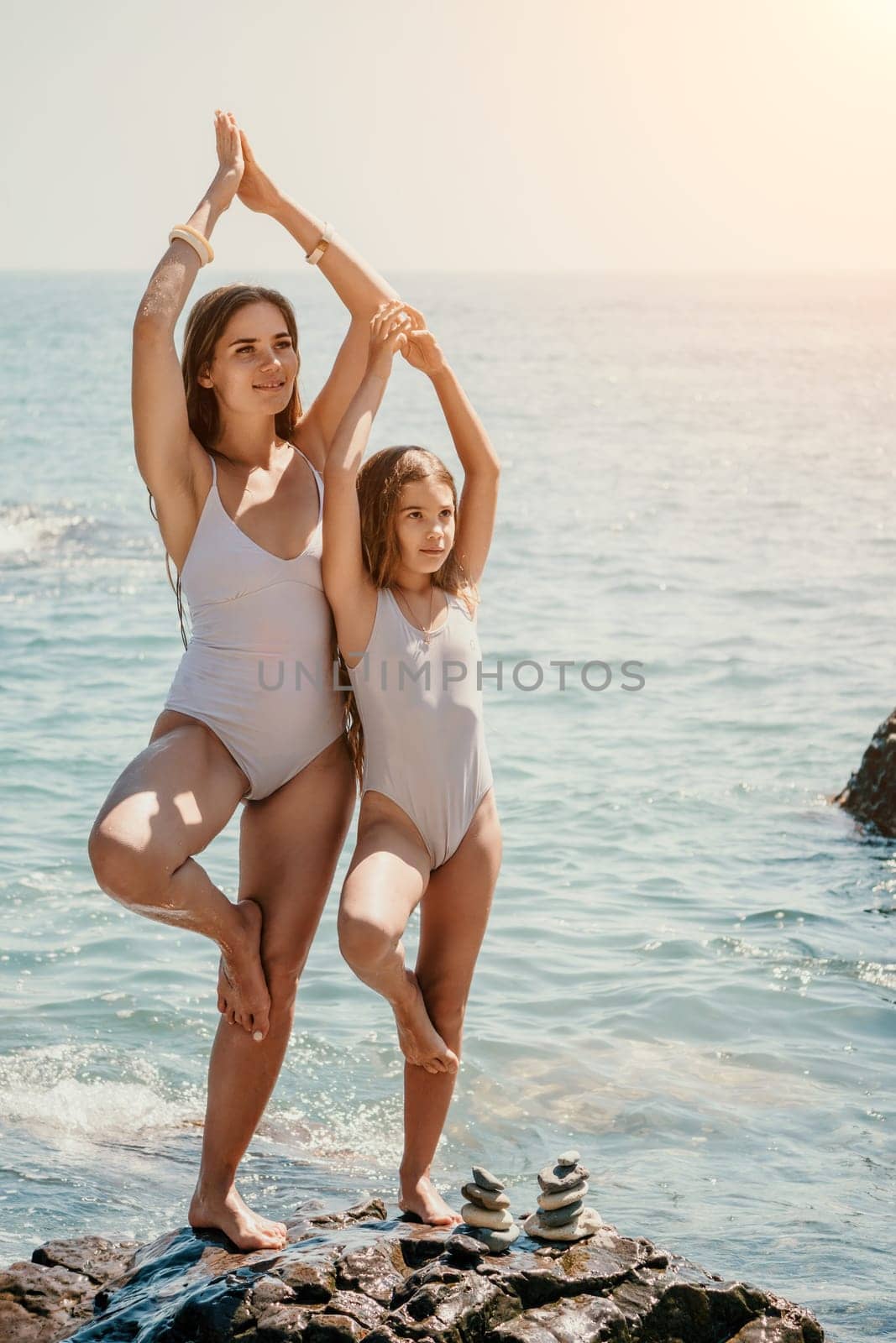 Woman and her daughter practicing balancing yoga pose on one leg up together on rock in the sea. Silhouette mother and daughter doing yoga at beach by panophotograph