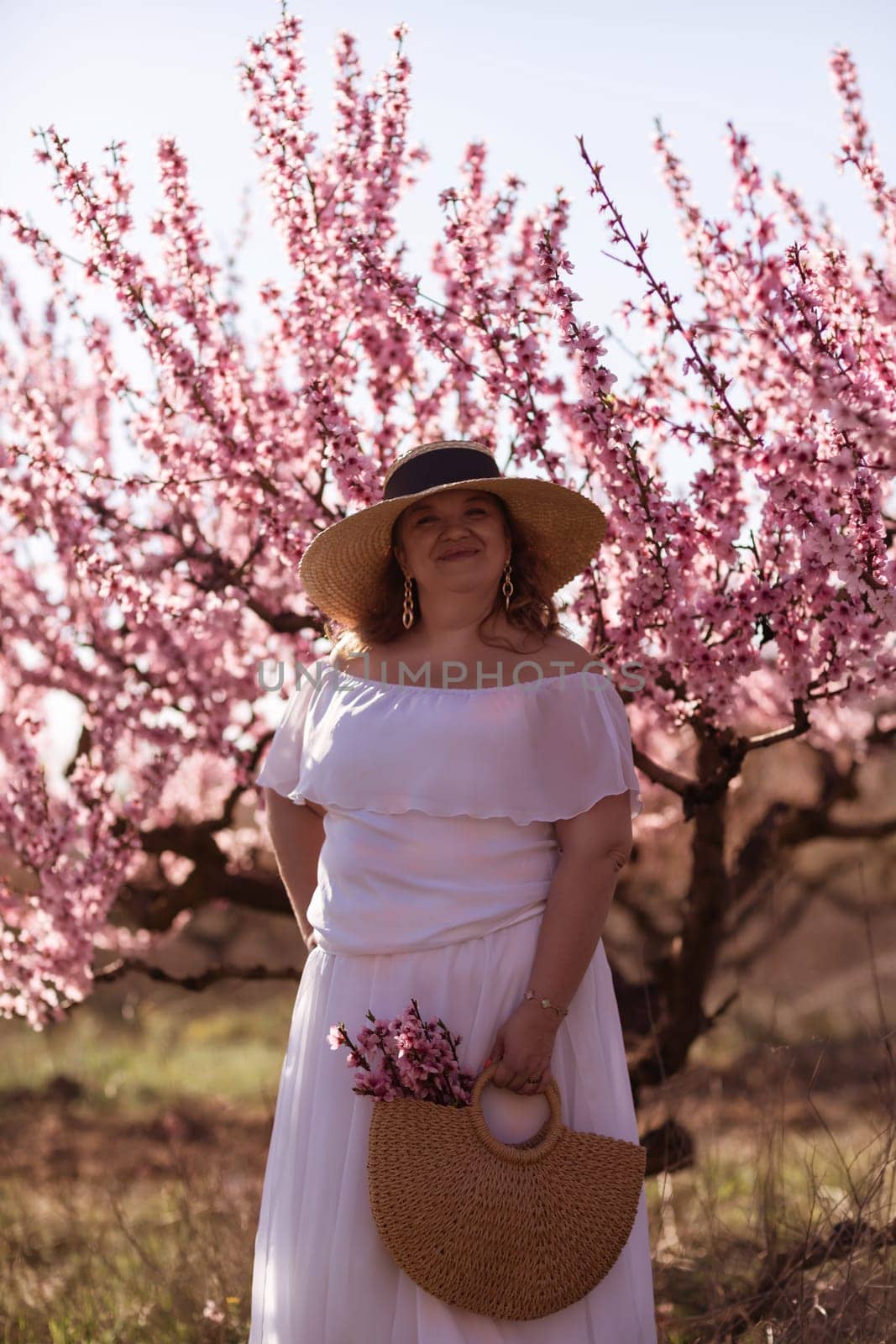 Woman blooming peach orchard. Against the backdrop of a picturesque peach orchard, a woman in a long white dress and hat enjoys a peaceful walk in the park, surrounded by the beauty of nature. by Matiunina