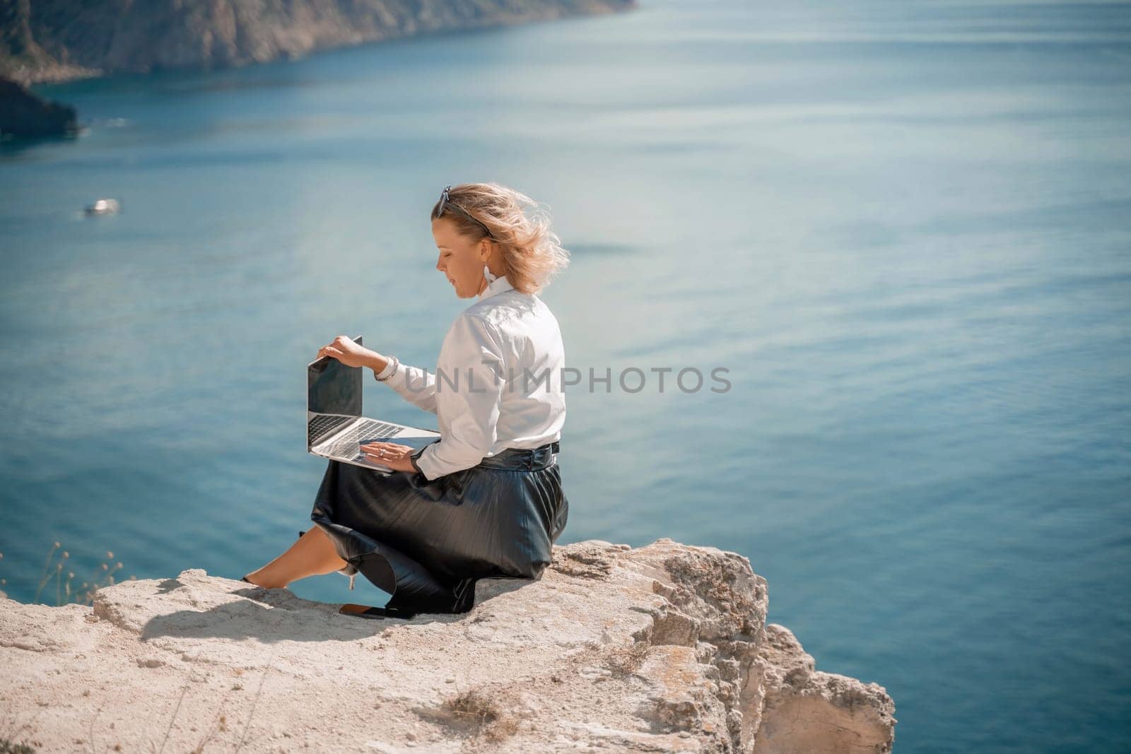 Business woman on nature in white shirt and black skirt. She works with an iPad in the open air with a beautiful view of the sea. The concept of remote work