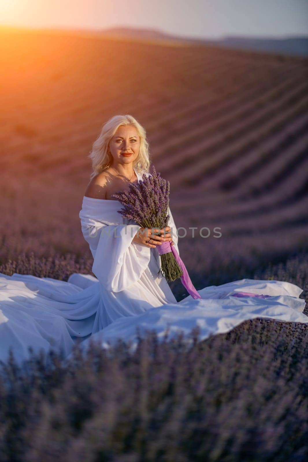 Blonde woman poses in lavender field at sunset. Happy woman in white dress holds lavender bouquet. Aromatherapy concept, lavender oil, photo session in lavender.