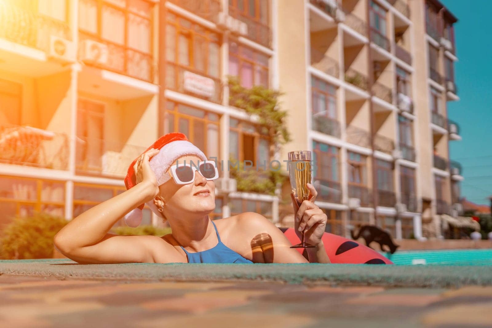 Woman pool Santa hat. A happy woman in a blue bikini, a red and white Santa hat and sunglasses poses near the pool with a glass of champagne standing nearby. Christmas holidays concept. by Matiunina