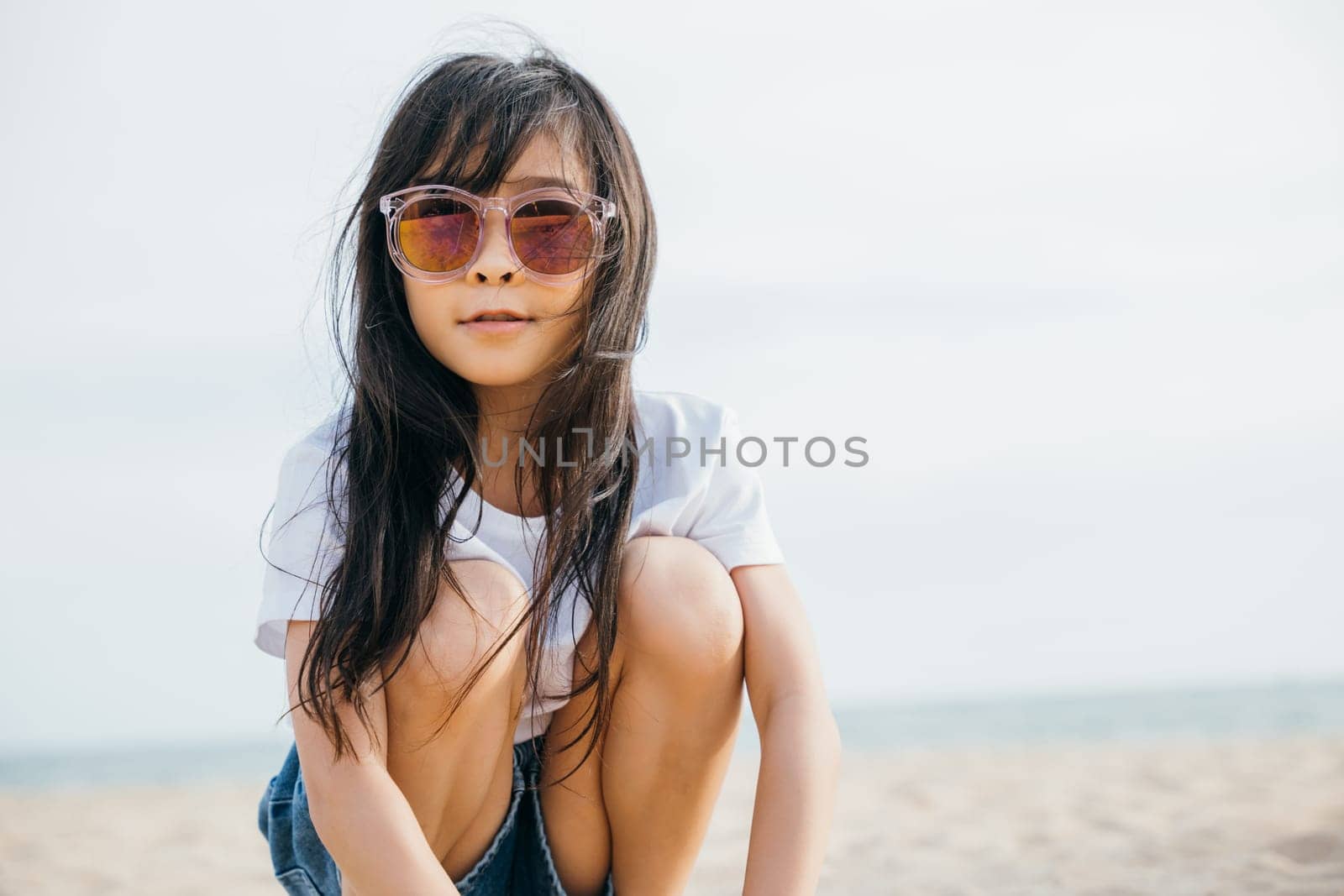 Beach style fashion, a child girl with trendy sunglasses sitting on a deck chair. Smiling portrait radiates joy and happiness. Windy hair stylish beachwear and carefree relaxation by Sorapop