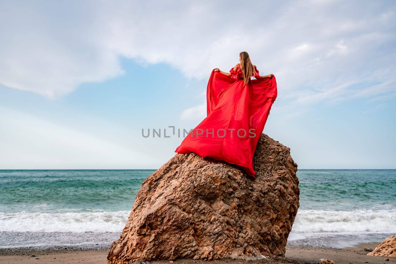 woman sea red dress. Woman with long hair on a sunny seashore in a red flowing dress, back view, silk fabric waving in the wind. Against the backdrop of the blue sky and mountains on the seashore