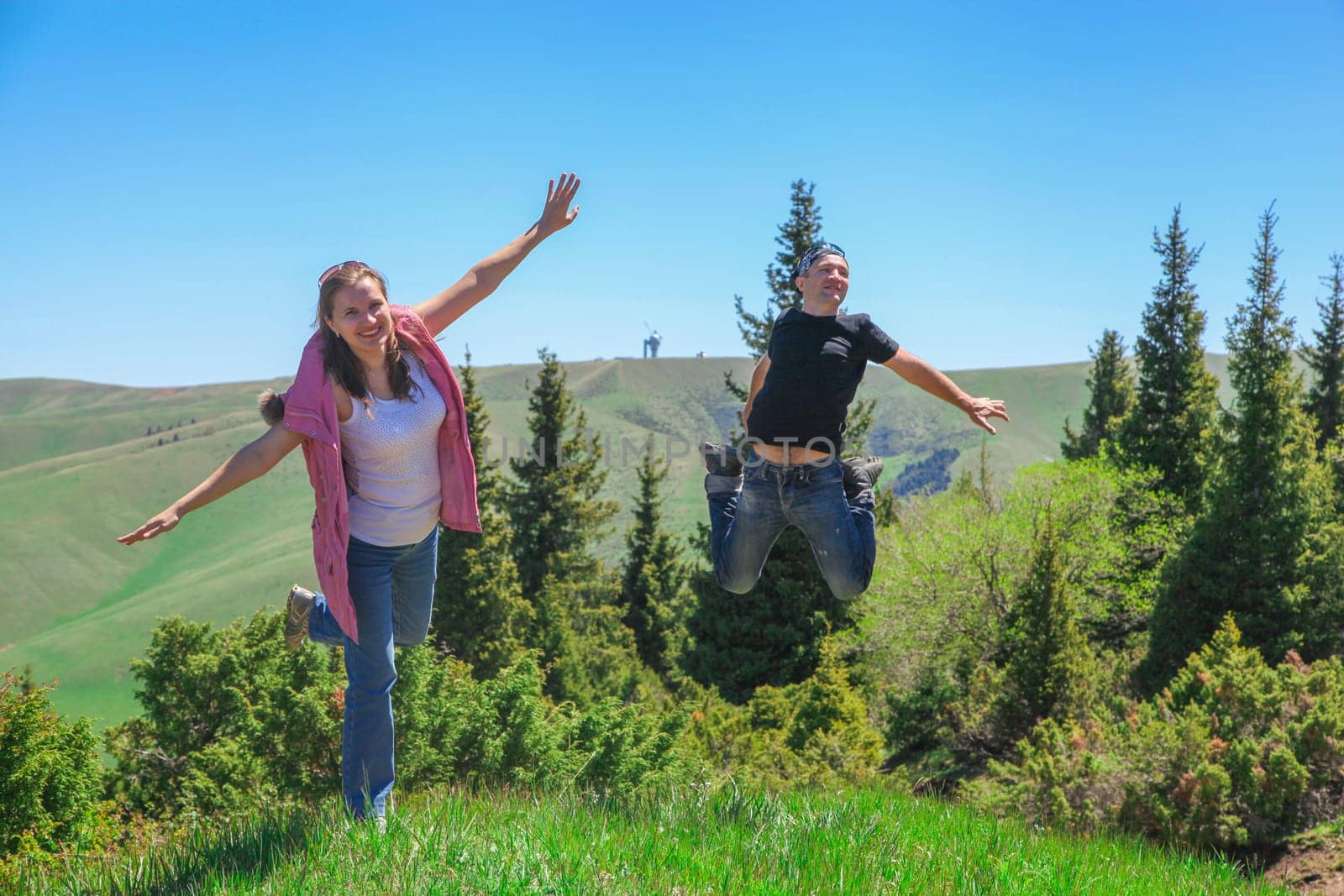 A happy couples. Woman in a pink waistcoat and blue jeans spread her arms and jumps on one foot. A man with a smiley face in black t-shirt and blue jeans jumps on both feet while spreading his arms