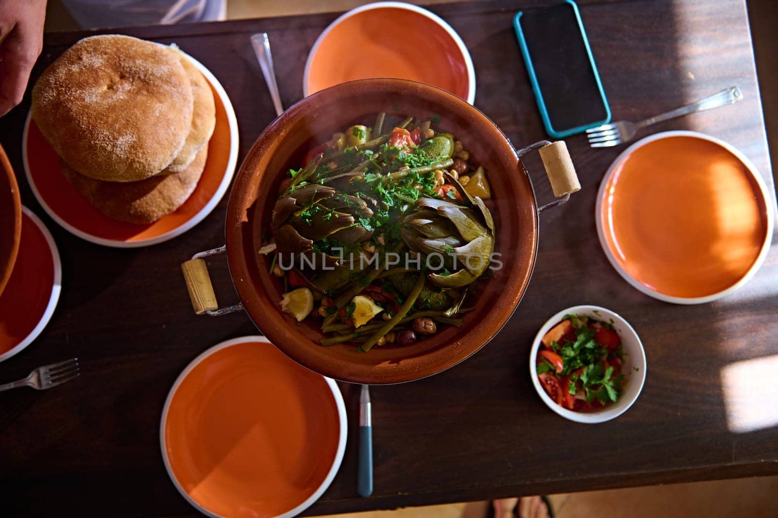Top view wooden table served with freshly backed traditional Moroccan wholegrain bread, a tagine with delicious organic veggies with steam. Background food and culinary blogs. Copy advertising space