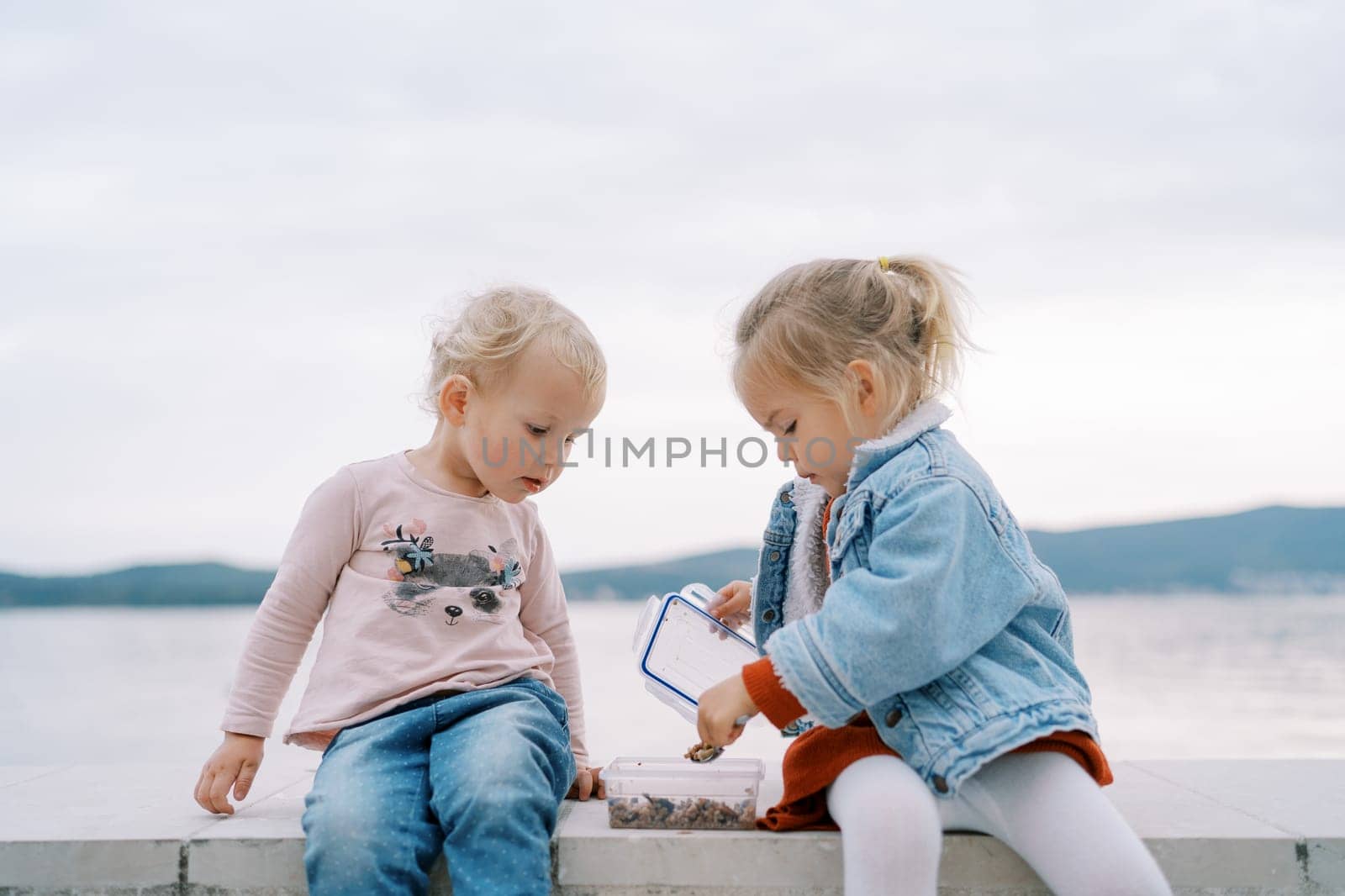 Little girl watches her friend open a lunchbox and scoop up granola with a spoon while sitting on a stone fence. High quality photo