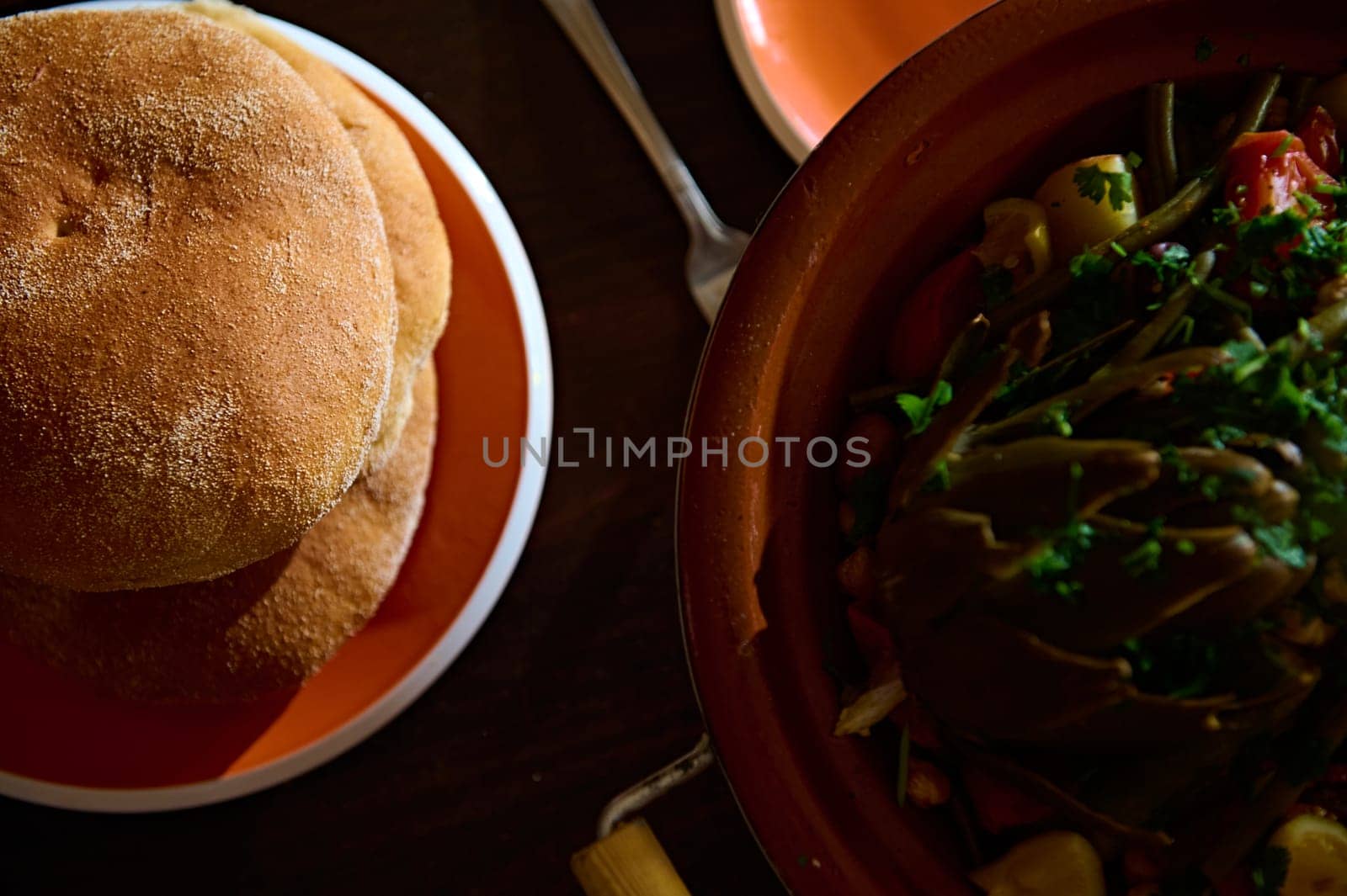 Top view of fresh baked wholegrain bread and tagine with steamed veggies on a wooden table. Close-up food photography for advertising and food blogs. Moroccan culinary, culture and traditional recipe