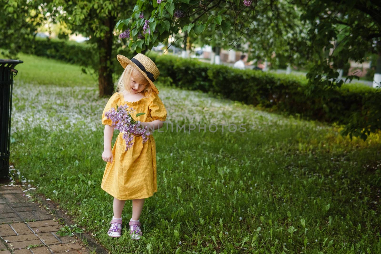 A little girl in a yellow dress and straw hat wearing a bouquet of lilacs. A walk in a spring park, blossoming lilacs