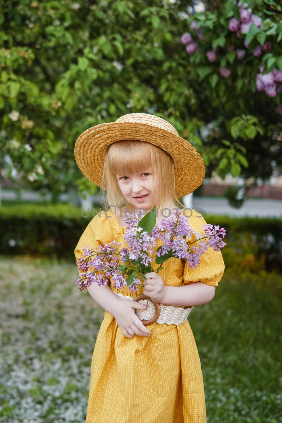 A little girl in a yellow dress and straw hat wearing a bouquet of lilacs. A walk in a spring park, blossoming lilacs