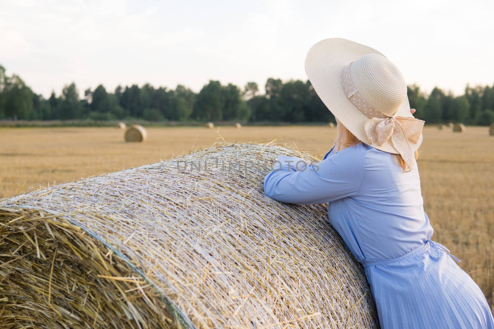 A red-haired woman in a hat and a blue dress walks in a field with haystacks