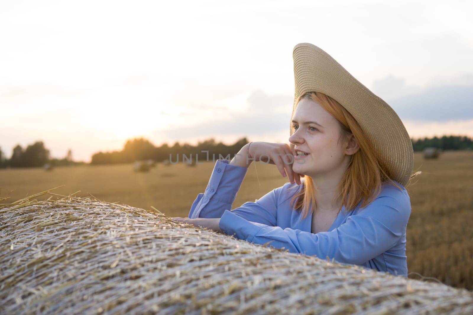 A red-haired woman in a hat and a blue dress walks in a field with haystacks