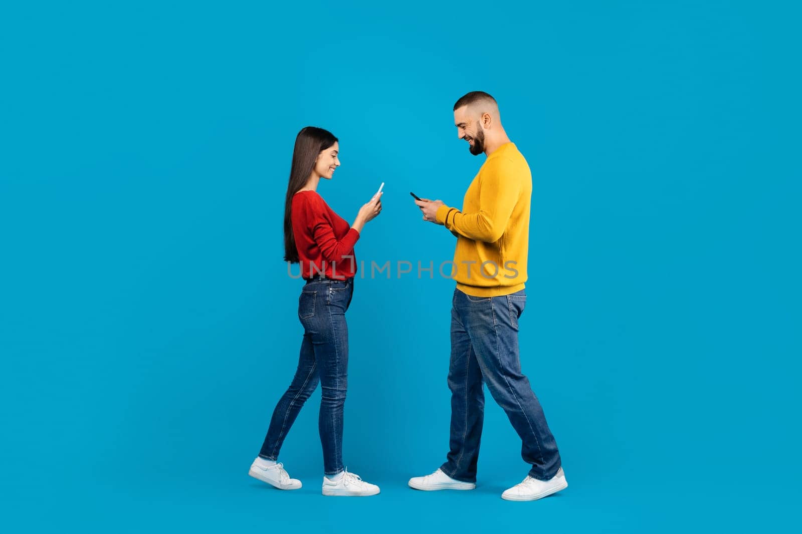 Communication, Technology And Lifestyle Concept. Smiling young couple using smartphones while walking past each other, man and woman standing isolated over blue studio background, full length