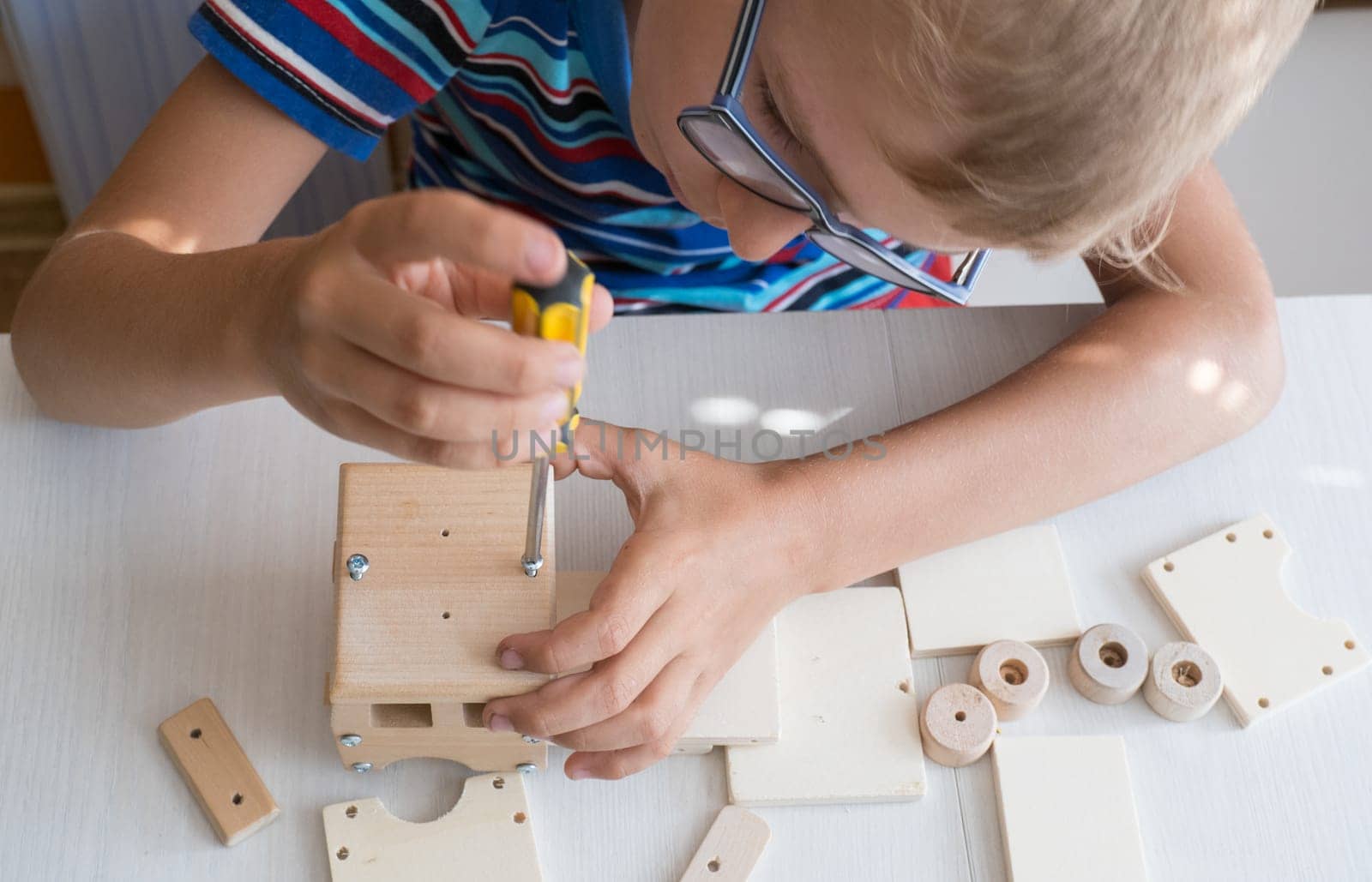 An 8-year-old boy assembles a wooden constructor by himself with a screwdriver. Close-up hands.