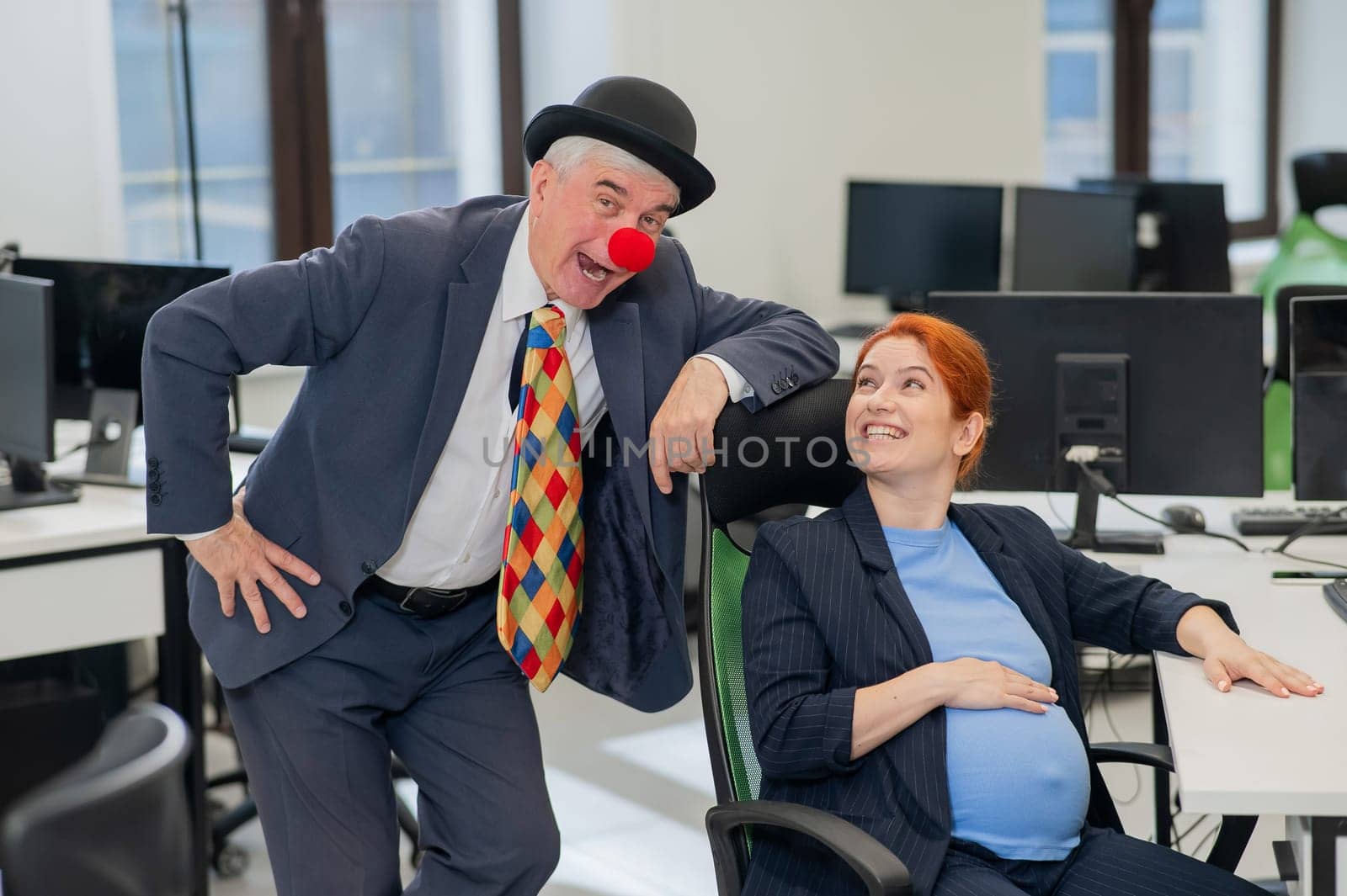 Elderly Caucasian man in clown costume amuses pregnant red-haired woman at work desk in office