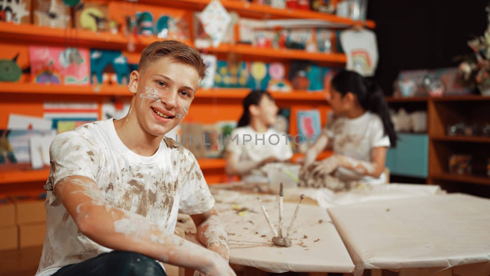 Highschool boy looking at camera while multicultural children modeling clay. Diverse student having pottery class together. Child smile while wearing white shirt with mud stained cloth. Edification.