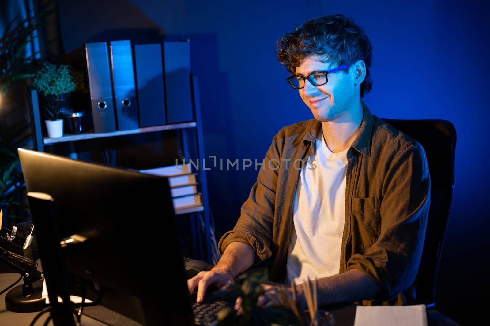 Smiling young businessman focusing on pc for creative startup project job at neon light modern office. Thinking marketing network online planning of creator to market channel design concept. Gusher.
