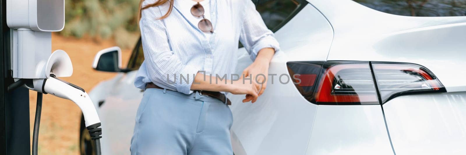 Panorama banner young woman recharging battery for electric car during autumnal road trip travel EV car in autumnal forest. Eco friendly travel on vacation during autumn with electric vehicle. Exalt