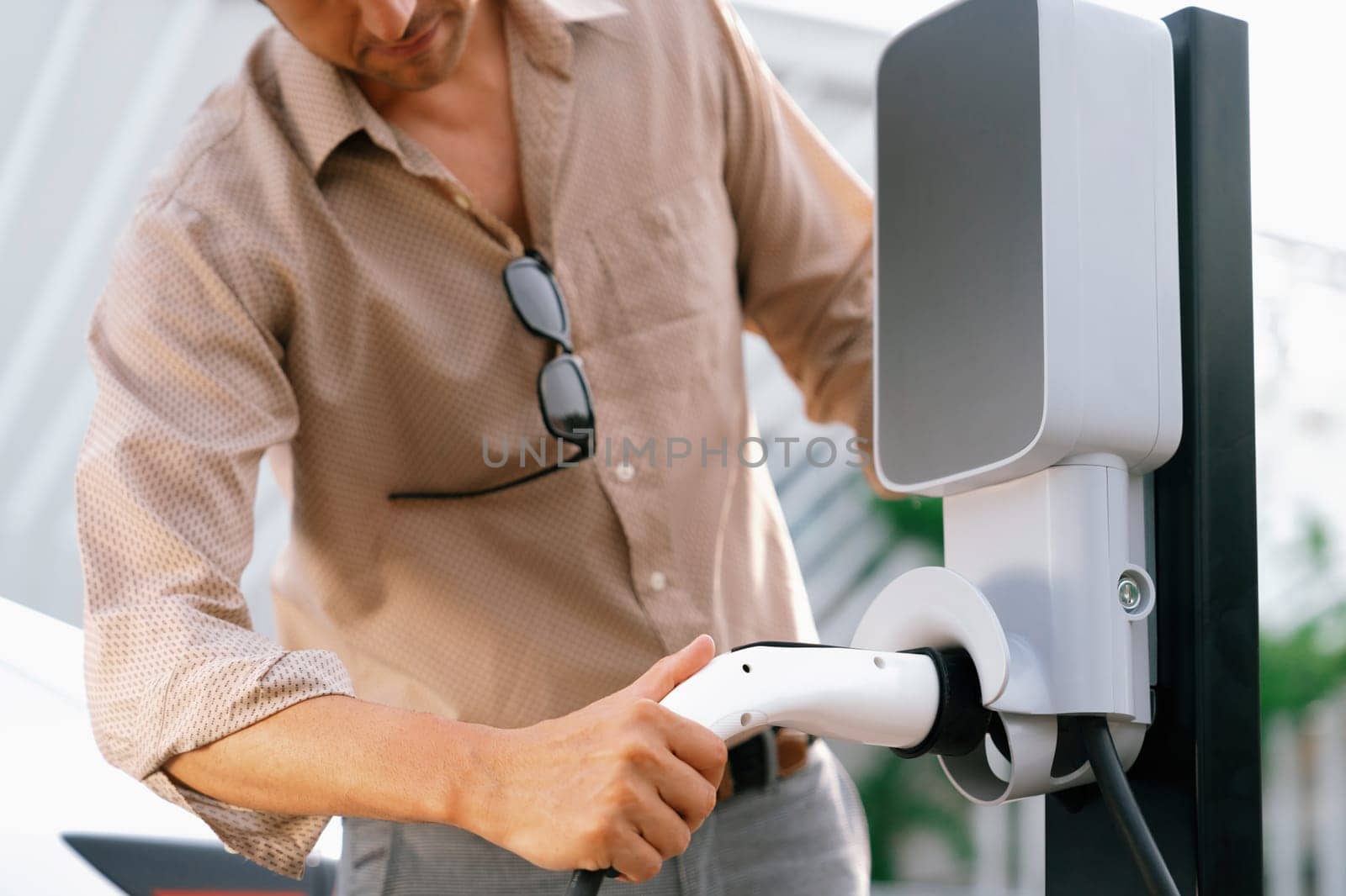 Young man put EV charger to recharge electric car's battery from charging station in city commercial parking lot. Rechargeable EV car for sustainable environmental friendly urban travel. Expedient