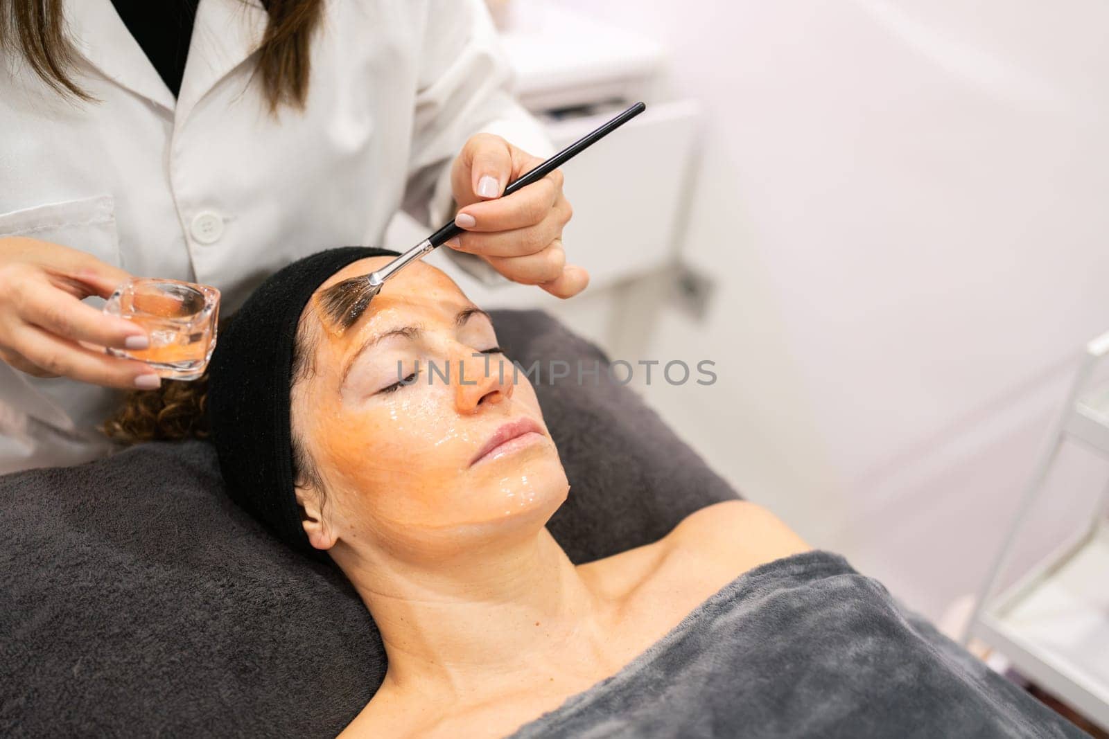 From above of crop anonymous cosmetician applying moisturizing product on skin of tranquil lady during beauty procedure in salon