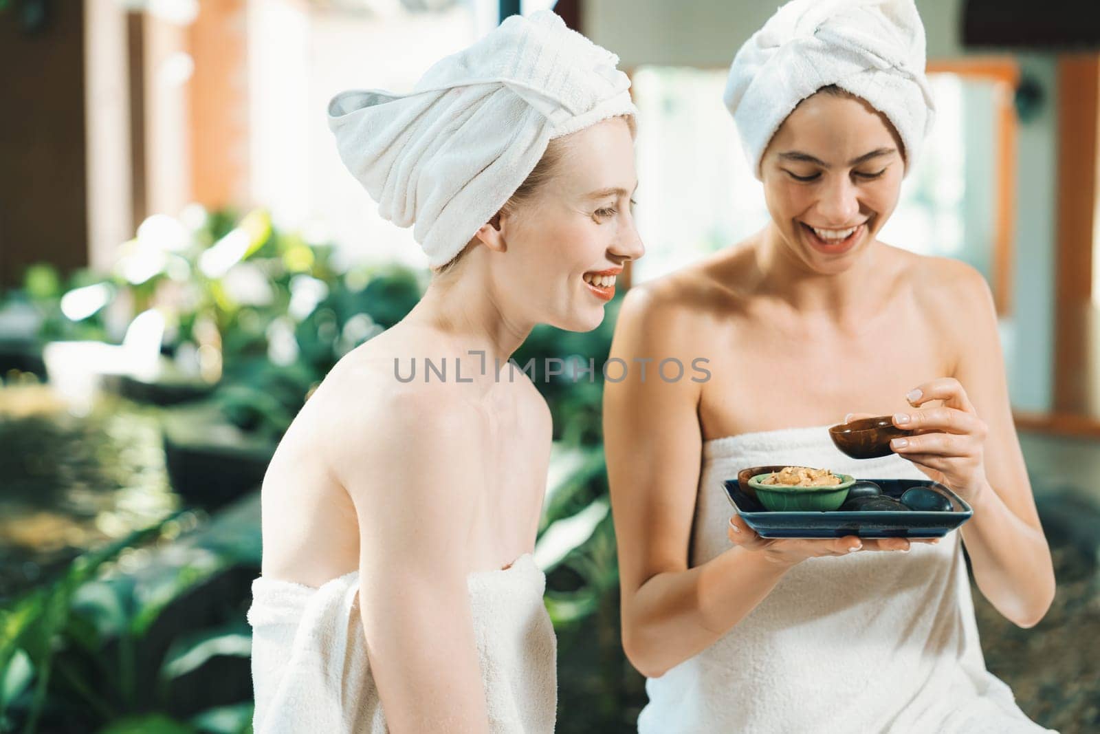 Couple of beautiful woman interested in homemade facial masks while sitting at spa salon. Attractive woman in white towel enjoy herbal masks with her friend surrounded by nature. Tranquility.