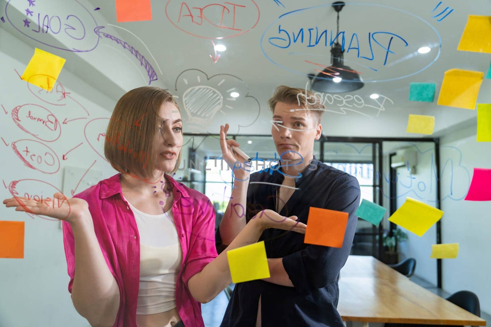 Couple of businesspeople discussing and brainstorming about solving marketing problem in front of glass board at business meeting by using mind map and sticky notes. Working together. Immaculate.