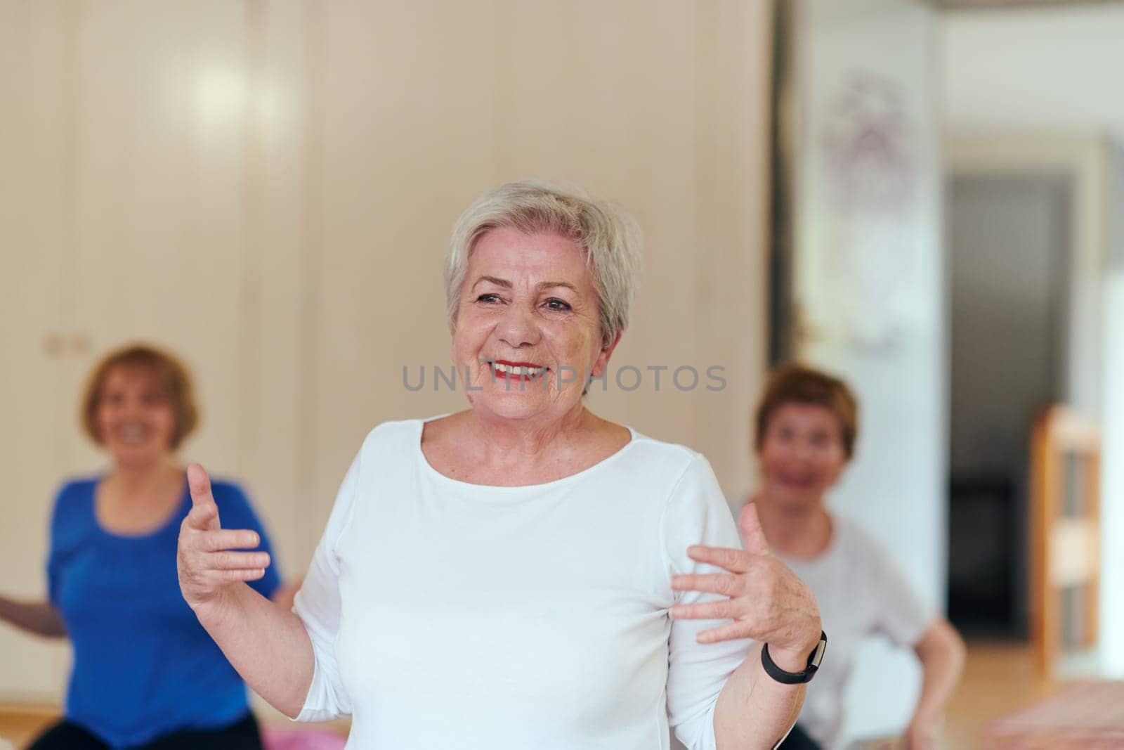 A group of senior women engage in various yoga exercises, including neck, back, and leg stretches, under the guidance of a trainer in a sunlit space, promoting well-being and harmony by dotshock