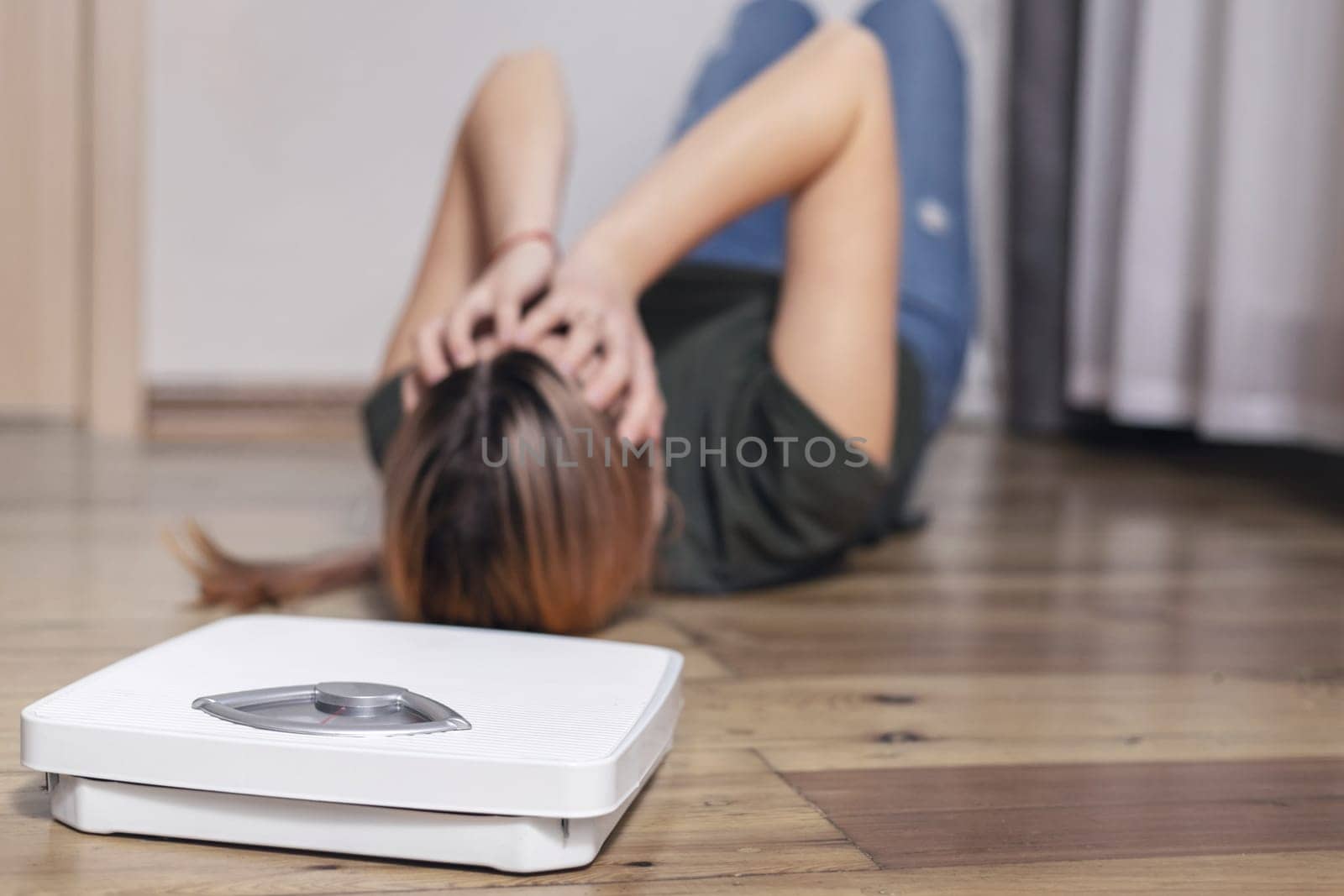 Scales and depressed young woman lying on the floor holding her head and covering her face with her hands.