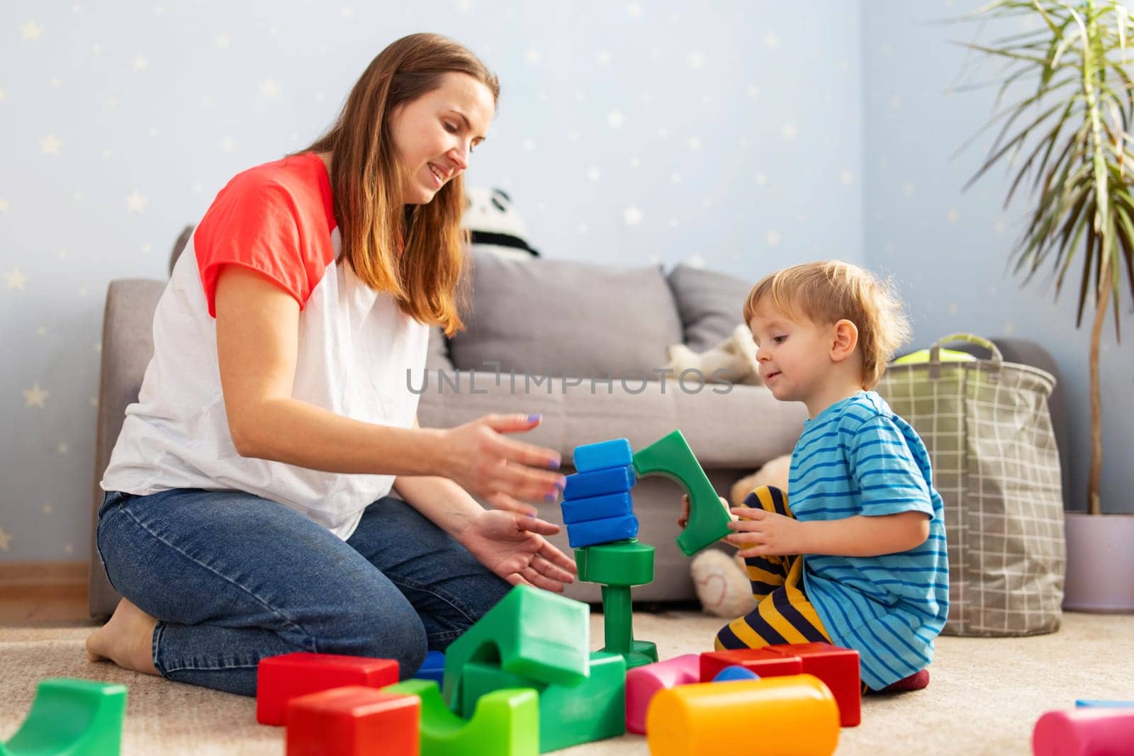 Cute little kid and child development specialist attractive young woman playing together with colorful blocks, sitting on the floor.