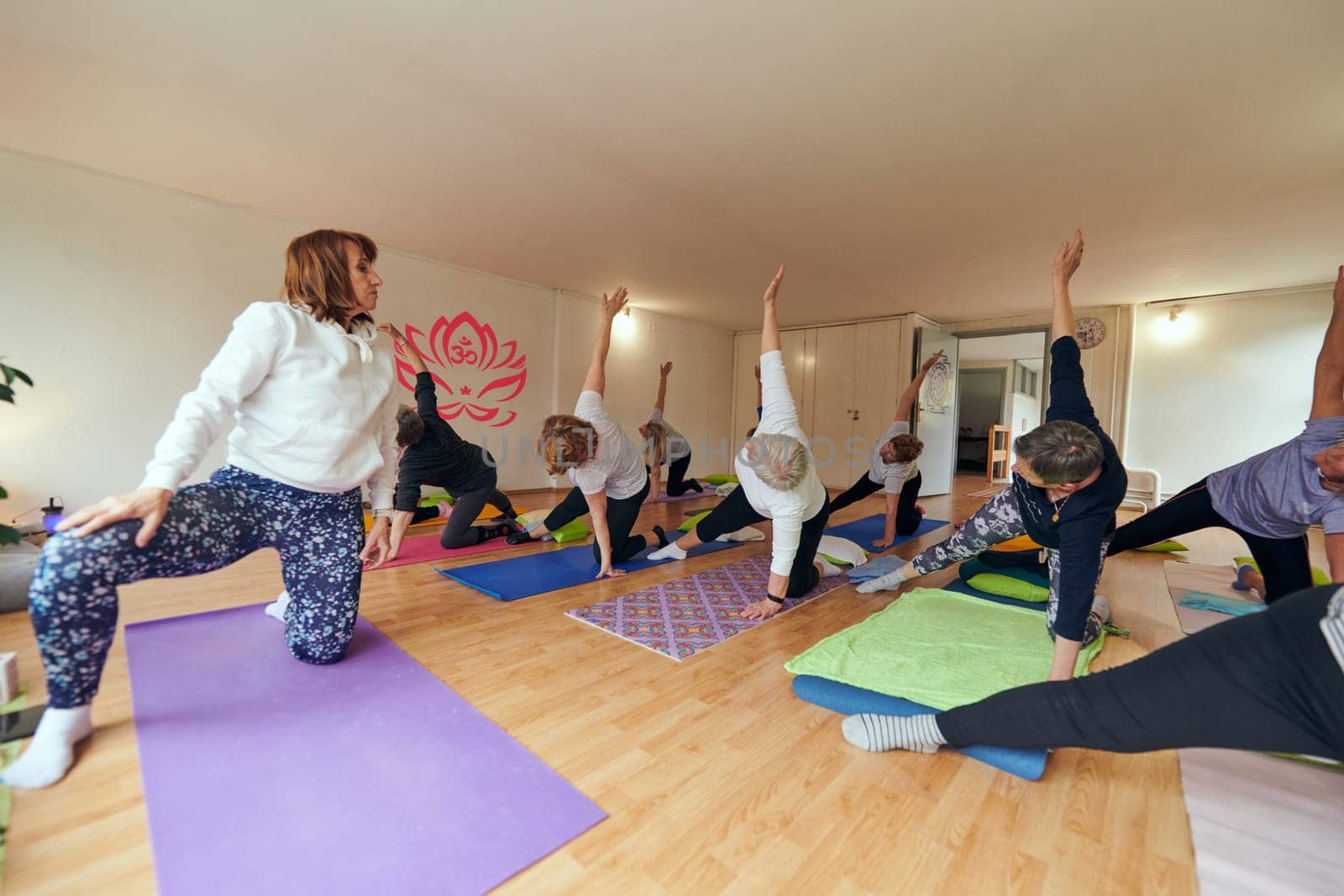 A group of senior women engage in various yoga exercises, including neck, back, and leg stretches, under the guidance of a trainer in a sunlit space, promoting well-being and harmony.