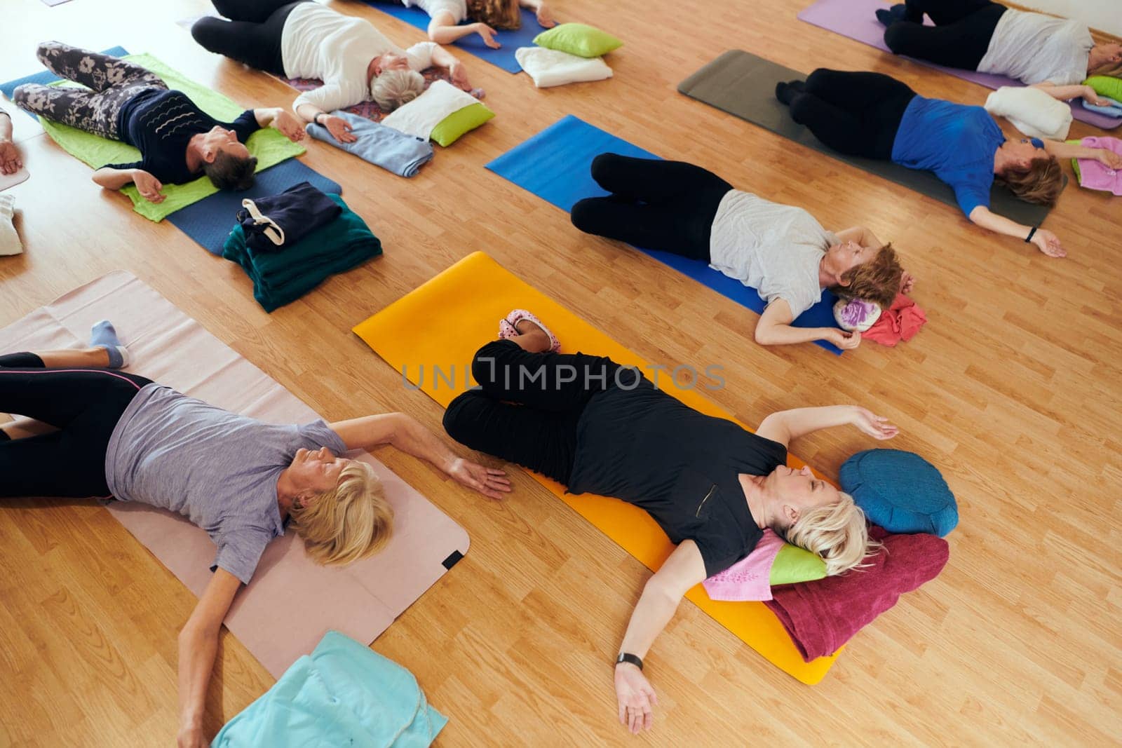 A group of senior women engage in various yoga exercises, including neck, back, and leg stretches, under the guidance of a trainer in a sunlit space, promoting well-being and harmony by dotshock