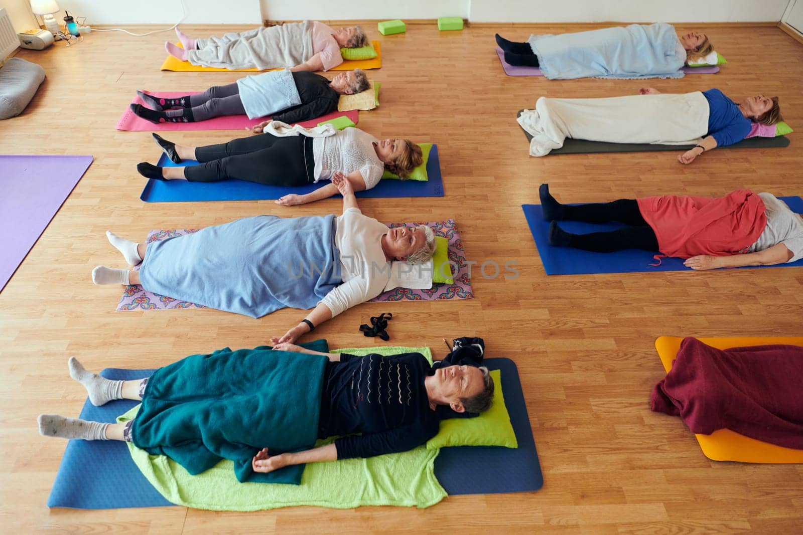 A group of senior women engage in various yoga exercises, including neck, back, and leg stretches, under the guidance of a trainer in a sunlit space, promoting well-being and harmony.