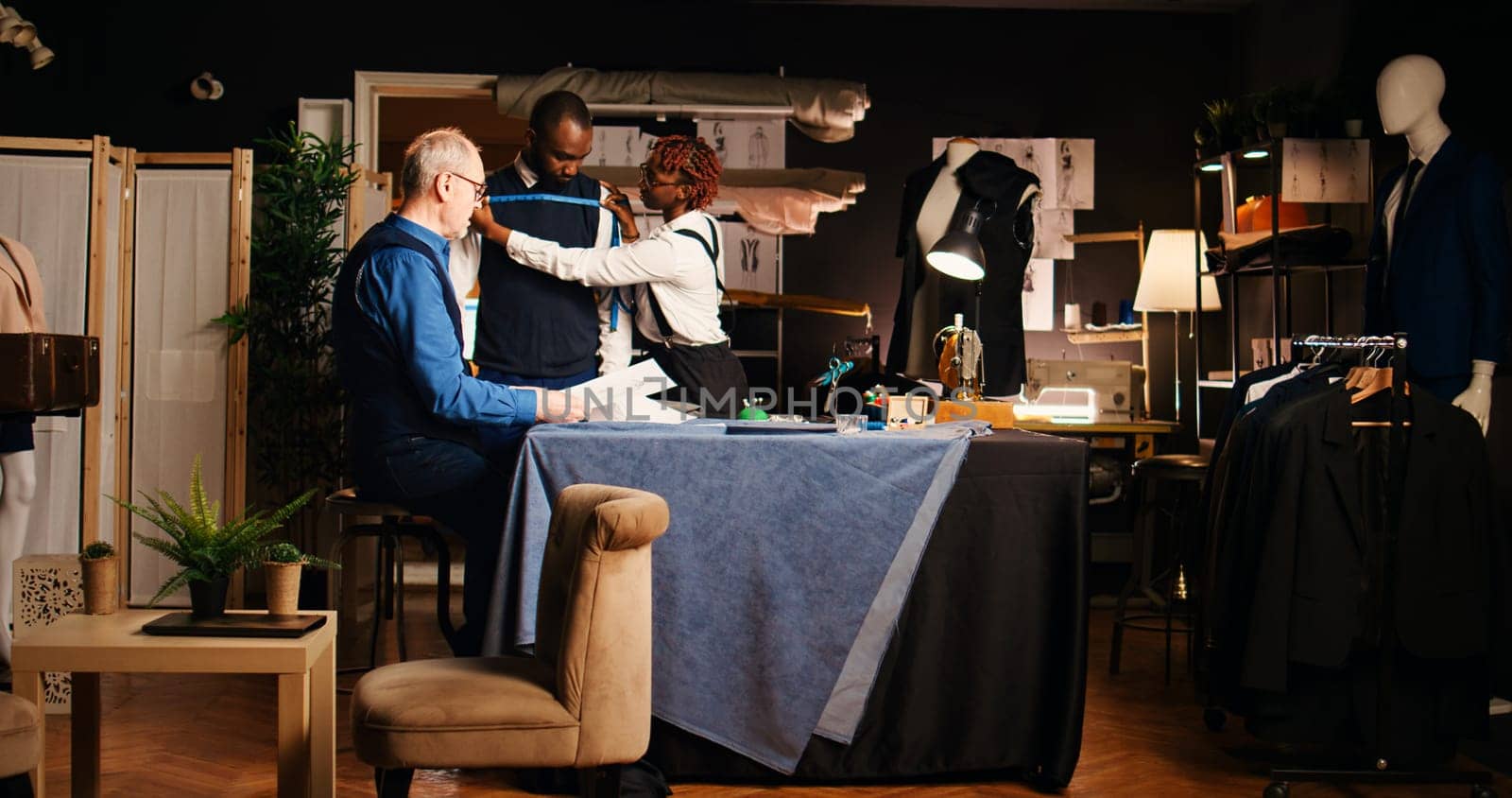 Customer working with senior tailor to design tailored suit in atelier, male client talking to couturier and employee about body sizes. Measuring tape and tailoring tools, small business.