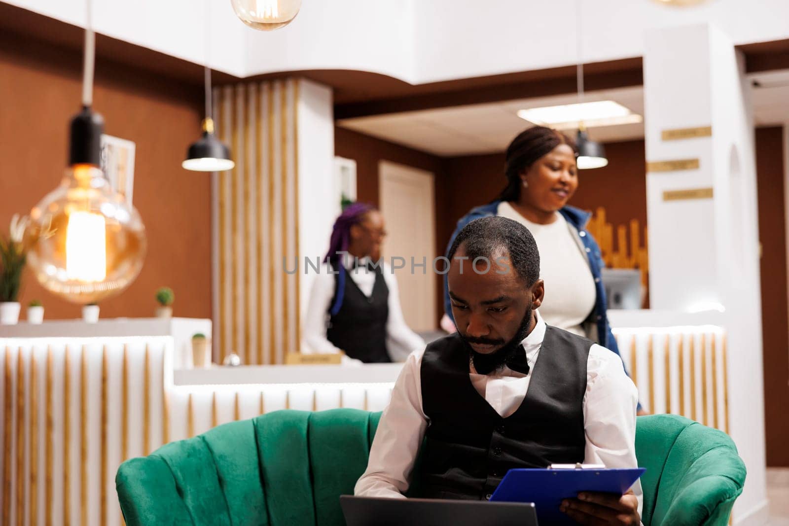 Focused African American man hospitality manager looking at computer pc screen while sitting in hotel lobby managing supplies. Black guy bellboy using laptop arranging transportation