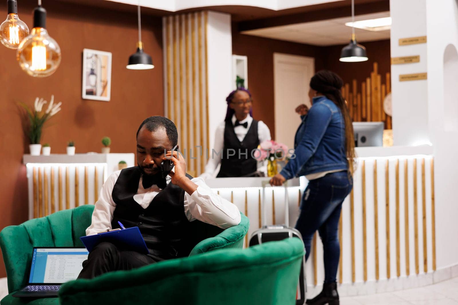 Young African American man hotel manager sitting in lobby talking by phone and taking notes, managing online bookings. Black guy bellboy holding smartphone ordering taxi for guest