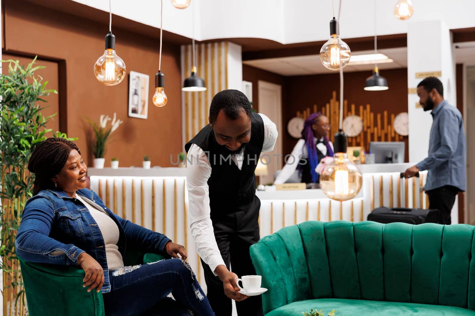 Happy smiling African American woman tourist relaxing in hotel lobby, looking at waiter serving coffee, feeling satisfied with customer service. Friendly restaurant worker putting cup on table