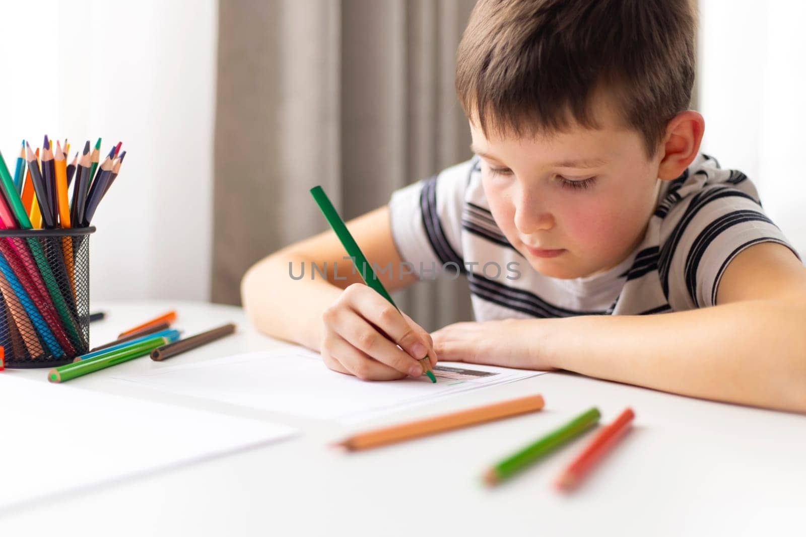 A child boy draws on white paper with colored pencils while sitting at a table.