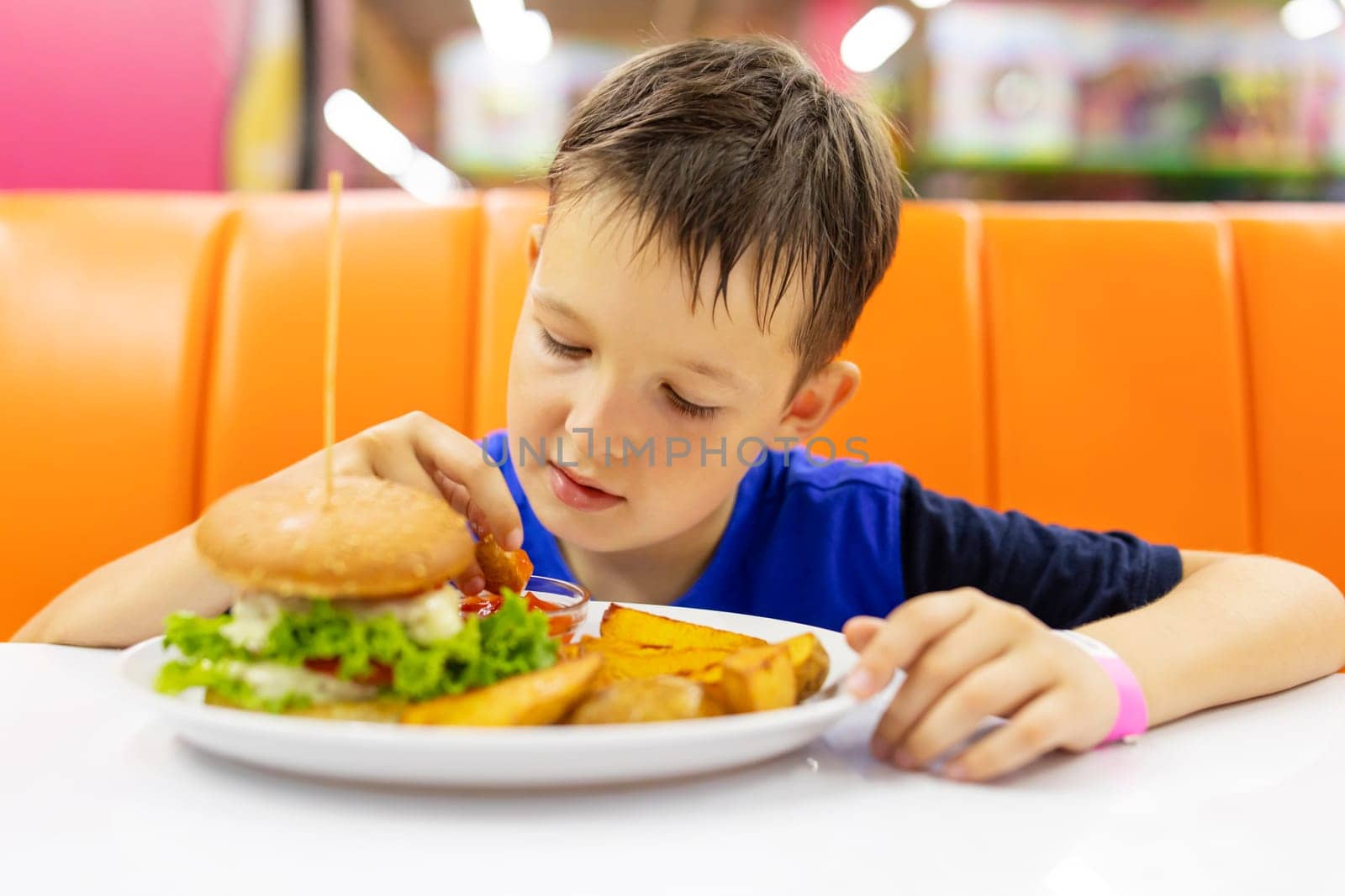 Child eating hamburger and French fries in a fast food restaurant. Child dipping a fried chip into tomato sauce