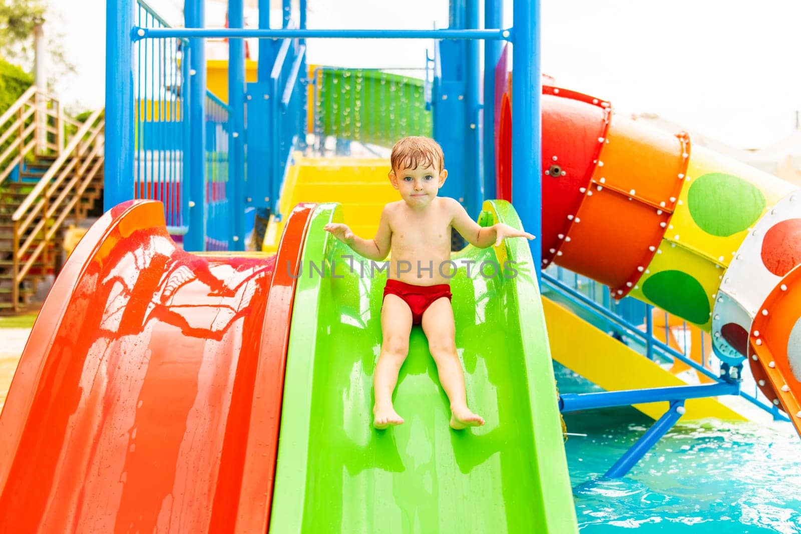 Happy laughing little boy playing on water slide in outdoor swimming pool on a hot summer day. Child sliding on aqua playground in tropical resort