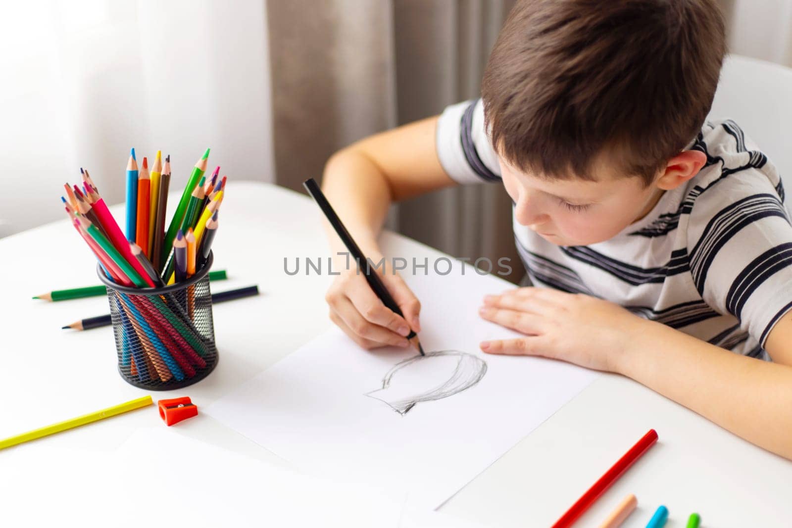 A child boy draws on white paper with colored pencils while sitting at a table.