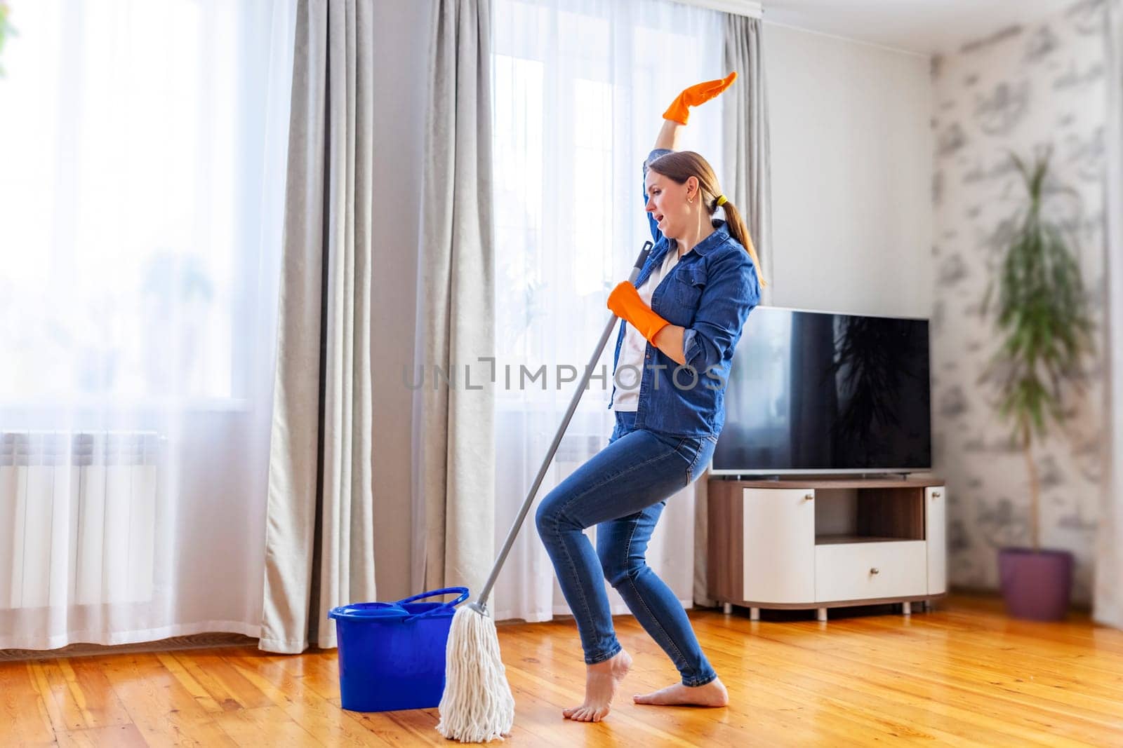 Funny happy young woman with mop singing, dancing and having fun while cleaning floor. Housewife enjoying domestic chores, doing home cleanup creatively