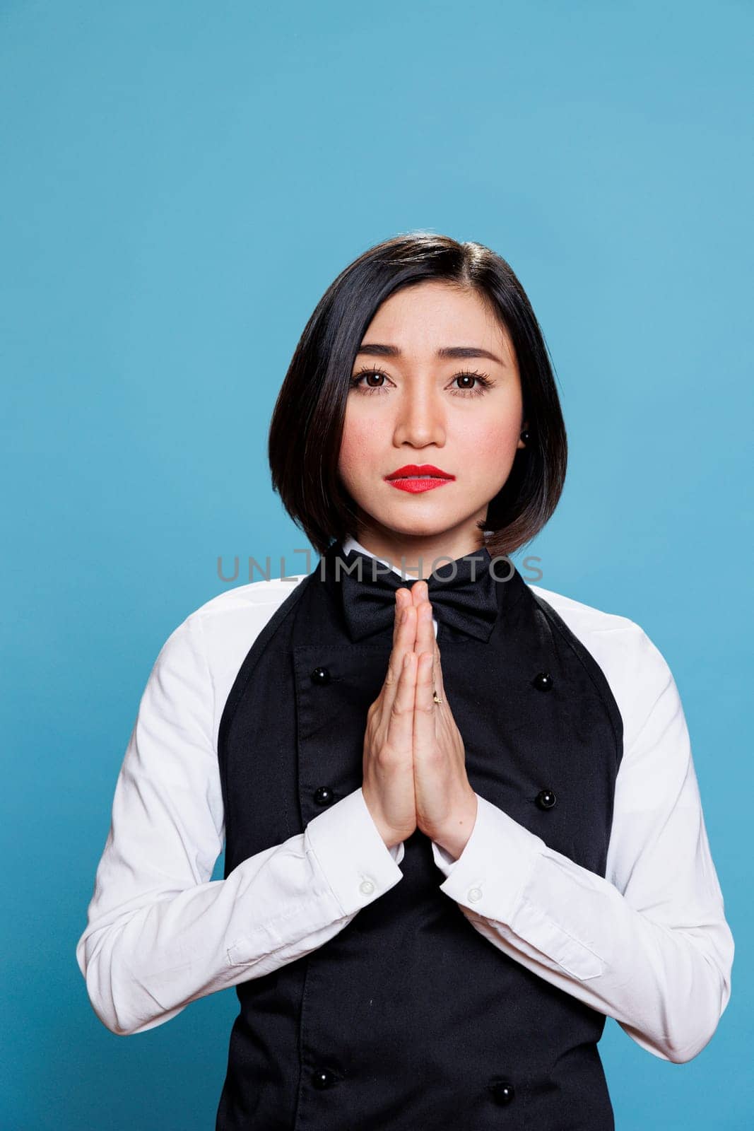 Young asian woman receptionist meditating with folded palms, relaxing and looking at camera. Attractive cafe waitress wearing uniform standing in zen pose portrait on blue background