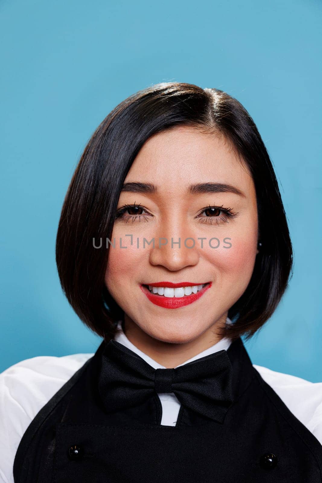 Smiling young attractive asian woman wearing waitress black and white uniform with bow tie closeup portrait. Cheerful woman receptionist looking at camera with positive facial expression