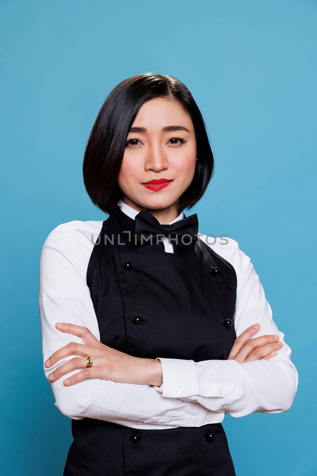 Restaurant woman receptionist wearing uniform smiling while posing with arms crossed portrait. Catering service asian waitress standing with folded hands and looking at camera with positive expression