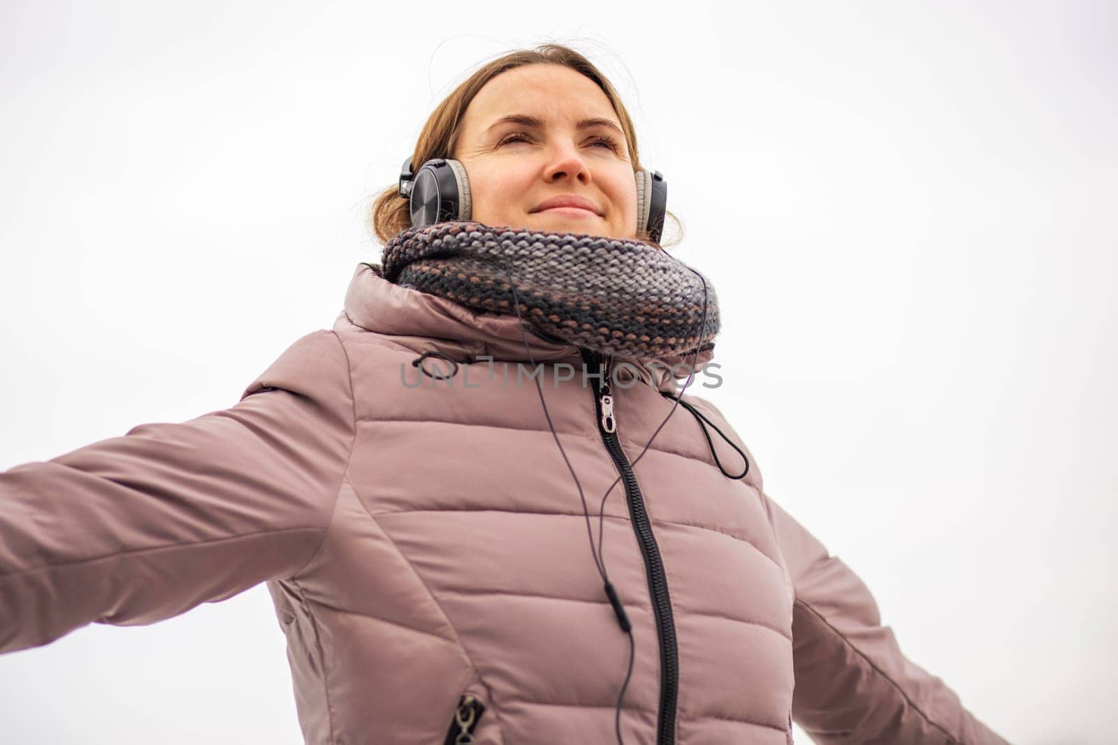 Joyful young woman in jacket listening to music via black headphones, breathing in the fresh air, relaxing in a park