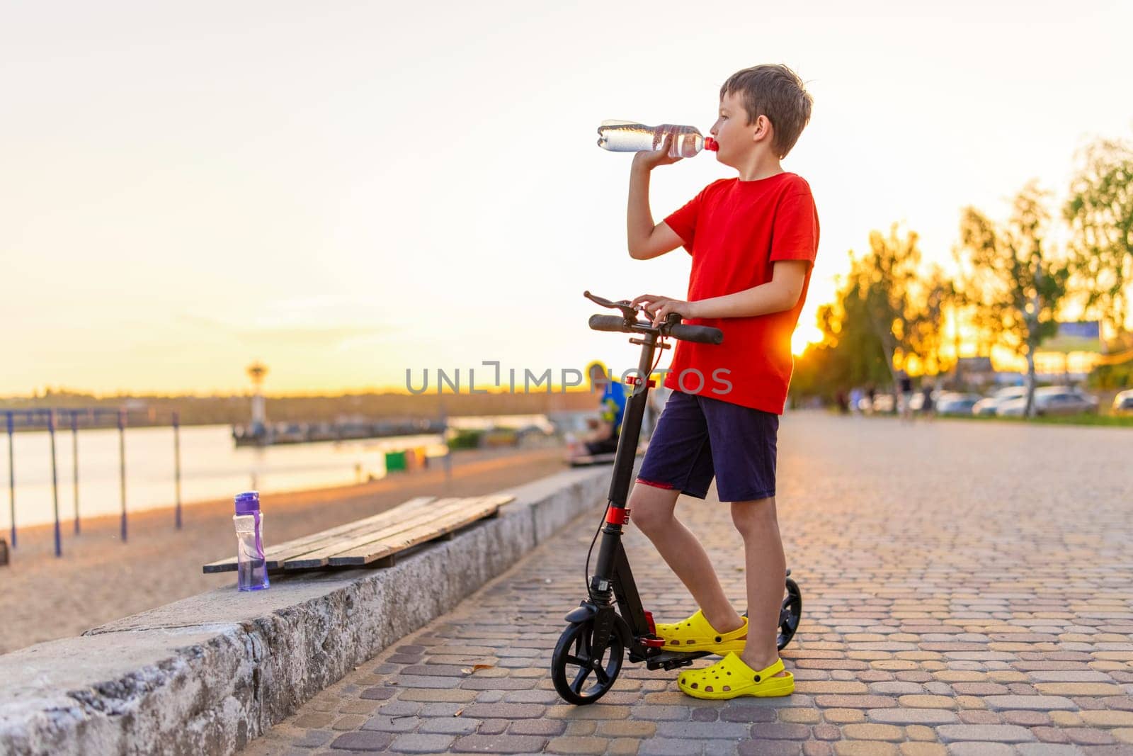 A boy drinks water from plastic bottle, standing with two-wheeled scooter. Child playing outdoors with scooters. Outdoor activities.