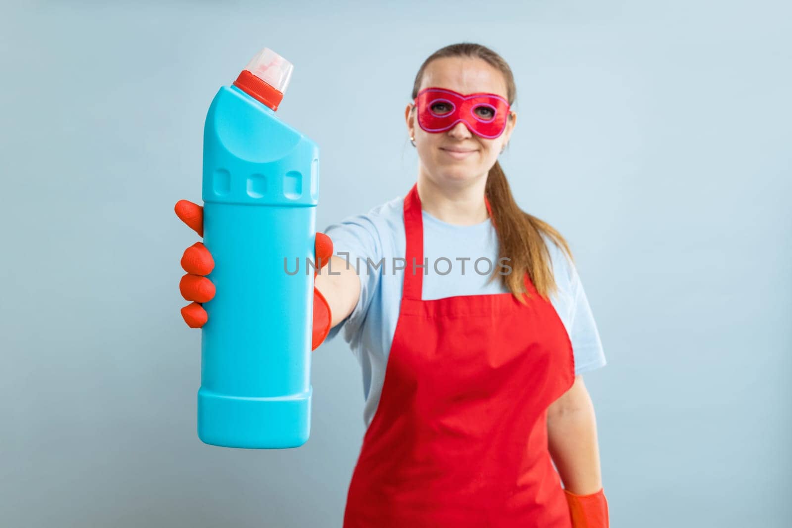Young woman in red mask, rubber gloves and apron holding detergent bottle on blue background. Advertising detergent