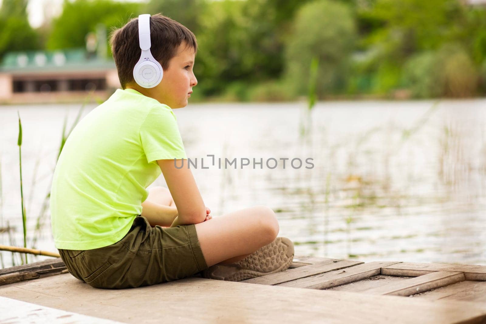 A boy with headphones listening to music at lakeside. Child sitting down by the lake listening to music